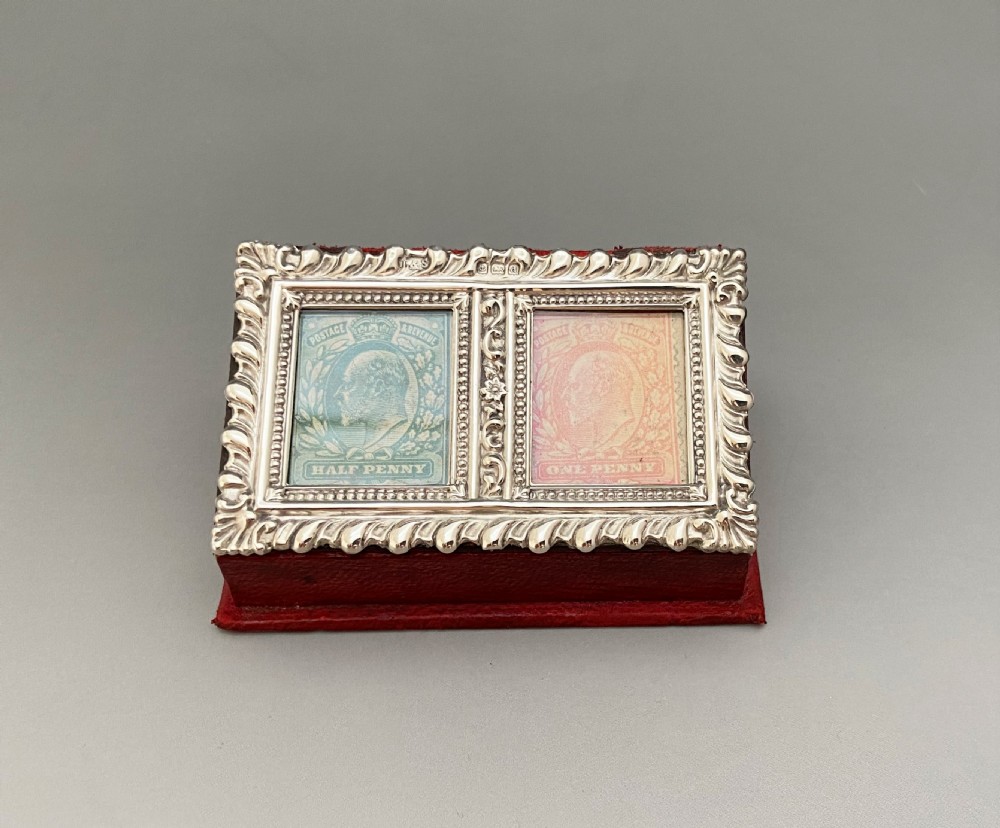An Edwardian sterling silver double stamp box featuring a decorative gadrooned border, two glazed top panels displaying Edward VII stamps, and a red leather base.