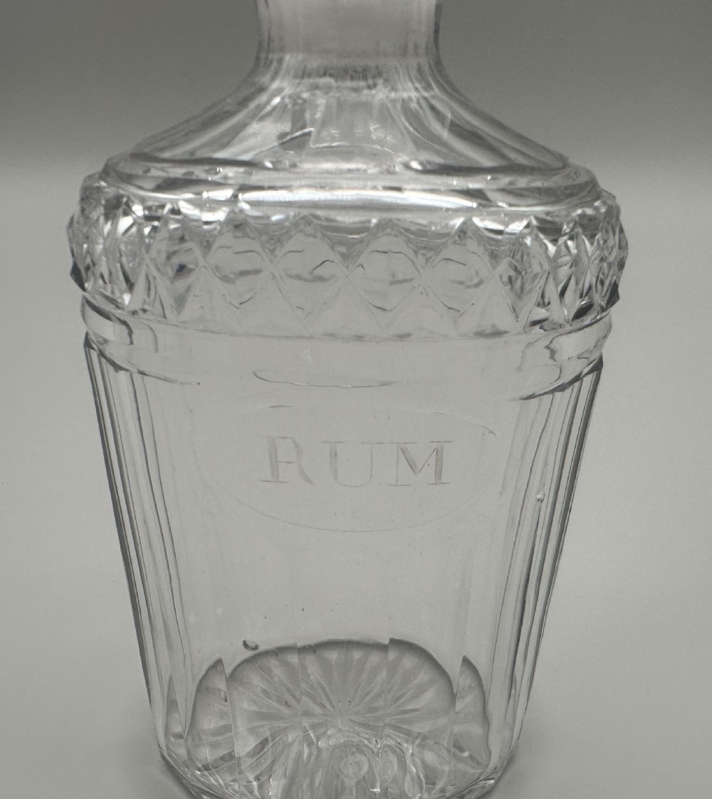 A clear Regency cut glass decanter featuring vertical fluting, a diamond-cut shoulder, an engraved oval cartouche reading RUM, and a fitted faceted stopper. View X