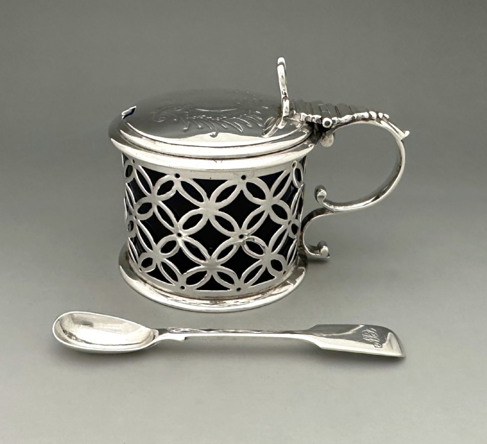A William IV sterling silver drum mustard pot featuring openwork geometric fretwork, a fitted blue glass liner, an engraved flat lid with a thumbpiece, a scroll handle, and a silver spoon.