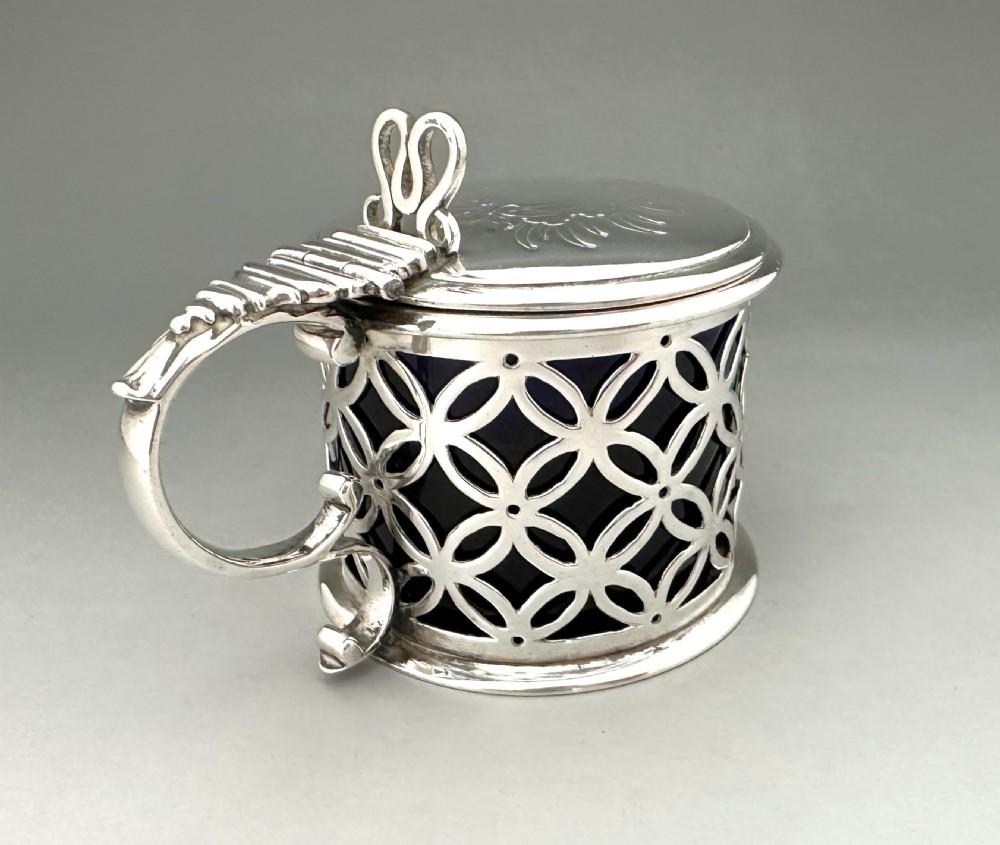 A William IV sterling silver drum mustard pot featuring openwork geometric fretwork, a fitted blue glass liner, an engraved flat lid with a thumbpiece, a scroll handle, and a silver spoon. View IV