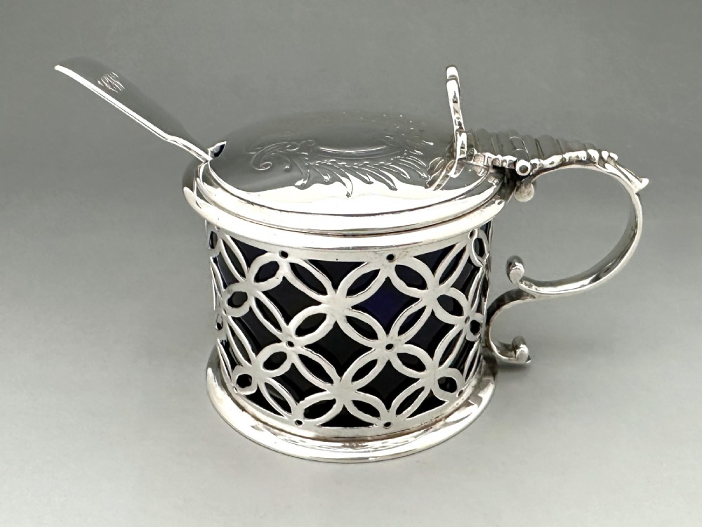 A William IV sterling silver drum mustard pot featuring openwork geometric fretwork, a fitted blue glass liner, an engraved flat lid with a thumbpiece, a scroll handle, and a silver spoon. View V
