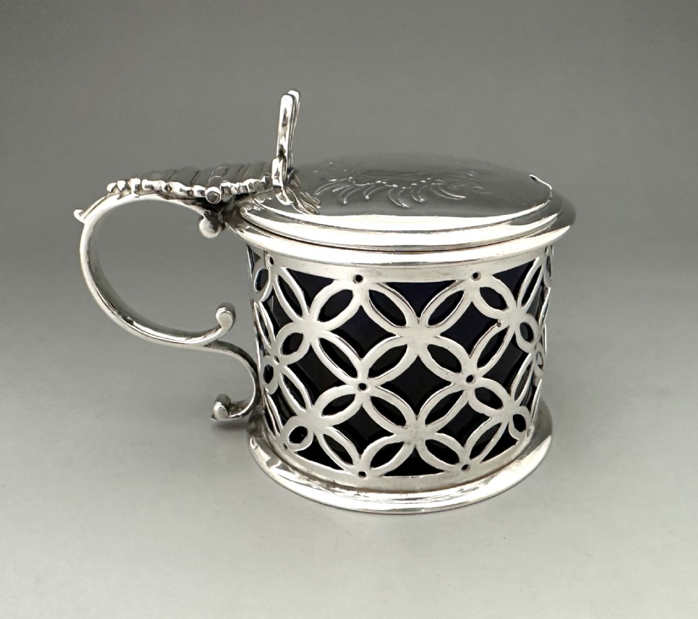 A William IV sterling silver drum mustard pot featuring openwork geometric fretwork, a fitted blue glass liner, an engraved flat lid with a thumbpiece, a scroll handle, and a silver spoon. View VI