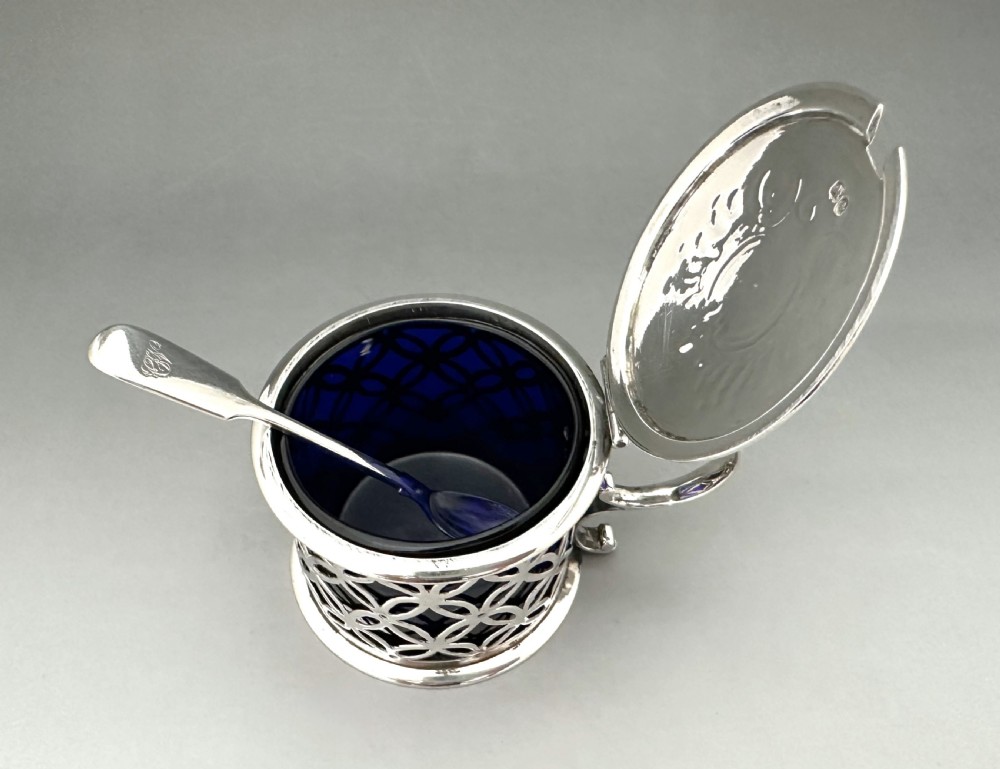 A William IV sterling silver drum mustard pot featuring openwork geometric fretwork, a fitted blue glass liner, an engraved flat lid with a thumbpiece, a scroll handle, and a silver spoon. View VII