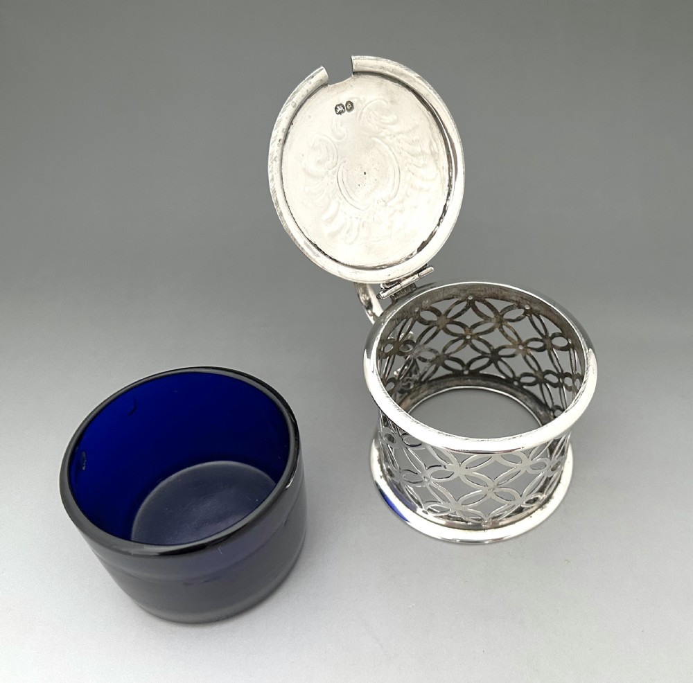 A William IV sterling silver drum mustard pot featuring openwork geometric fretwork, a fitted blue glass liner, an engraved flat lid with a thumbpiece, a scroll handle, and a silver spoon. View VIII