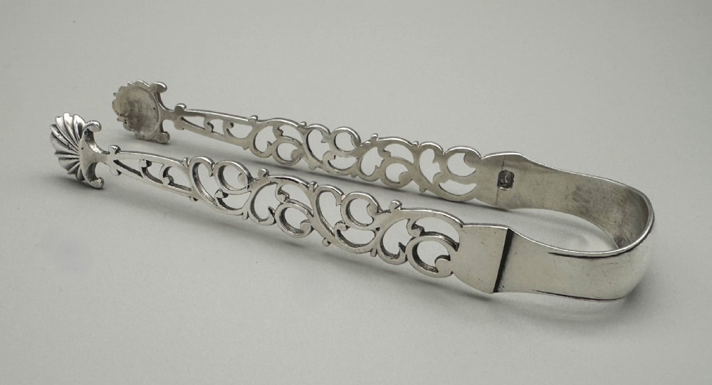 A pair of mid-18th-century cast silver sugar tongs with detailed pierced scrollwork arms and shell-shaped grips resting on a neutral background.
