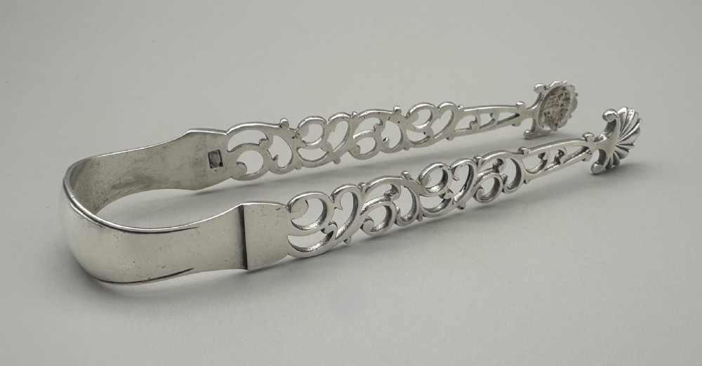 A pair of mid-18th-century cast silver sugar tongs with detailed pierced scrollwork arms and shell-shaped grips resting on a neutral background. View II