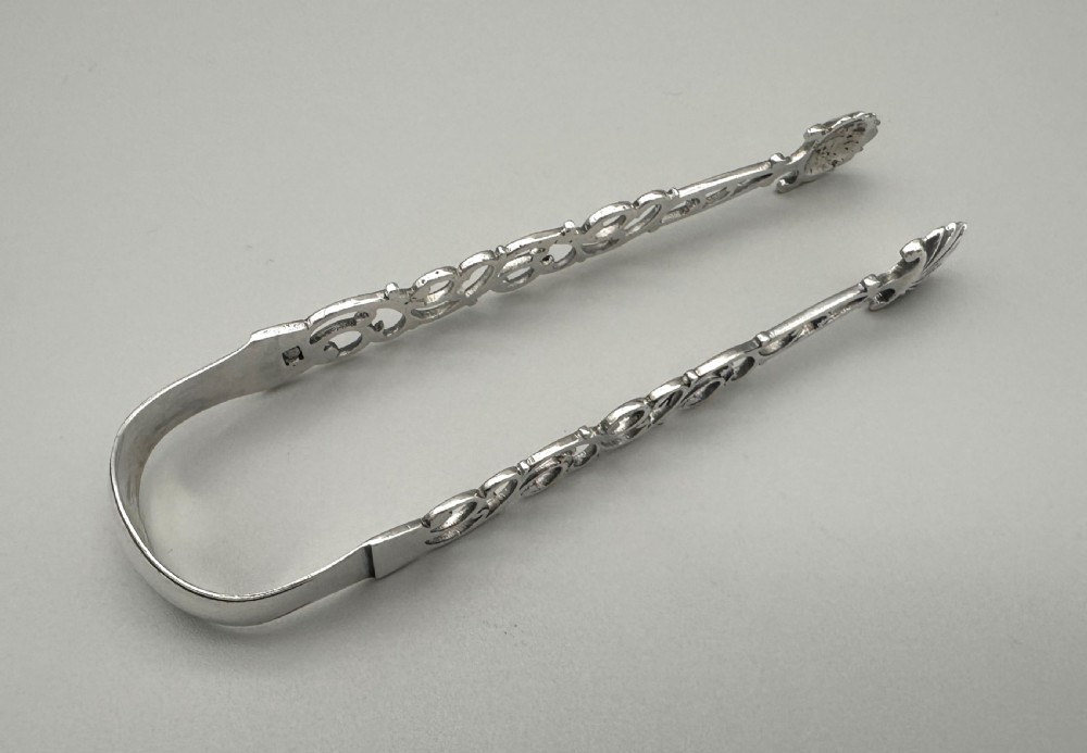 A pair of mid-18th-century cast silver sugar tongs with detailed pierced scrollwork arms and shell-shaped grips resting on a neutral background. View III