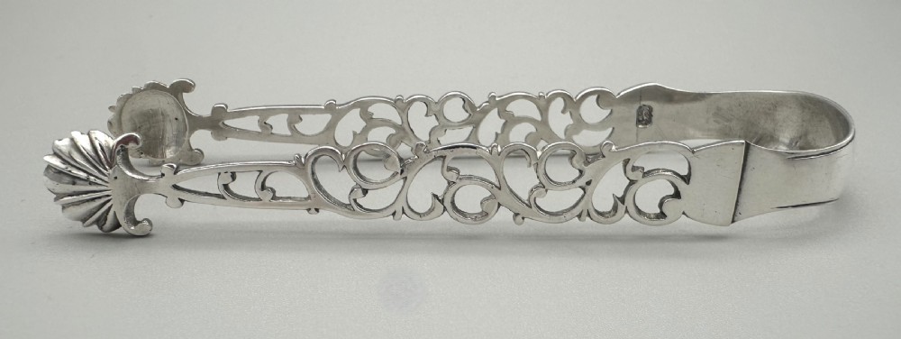 A pair of mid-18th-century cast silver sugar tongs with detailed pierced scrollwork arms and shell-shaped grips resting on a neutral background. View IV