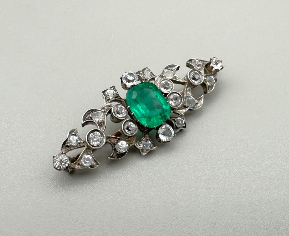 An antique French elongated brooch crafted from silver-topped gold, featuring a central cushion-cut green emerald surrounded by pierced scrollwork set with bright stones.