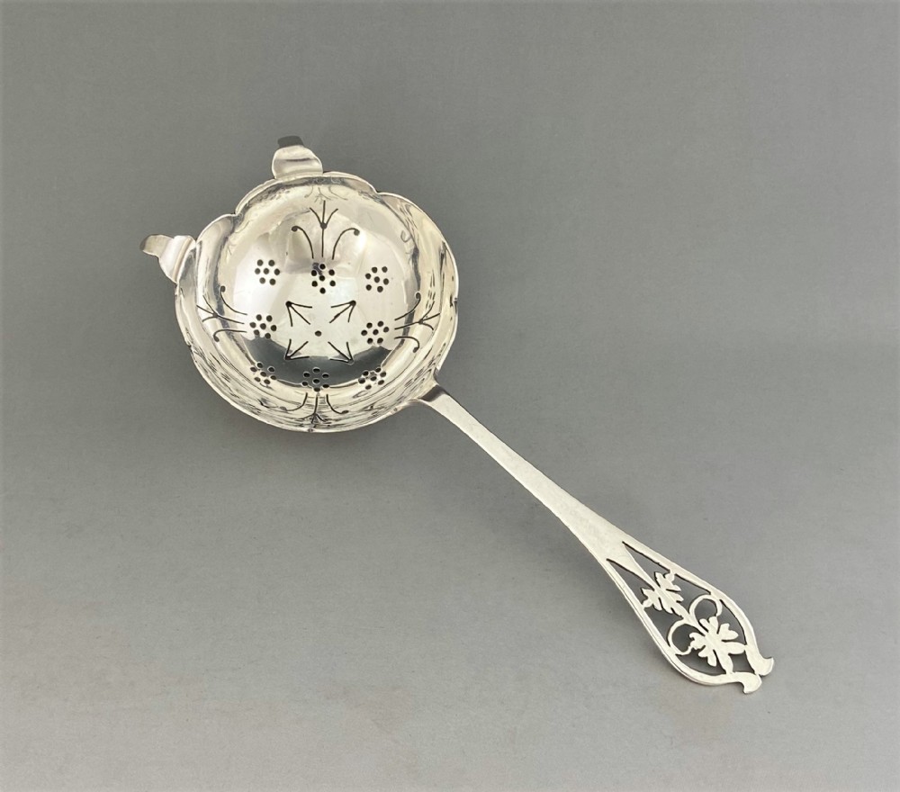 A silver tea strainer featuring a pierced bowl with geometric drainage holes and a decoratively cut-out flat handle.
