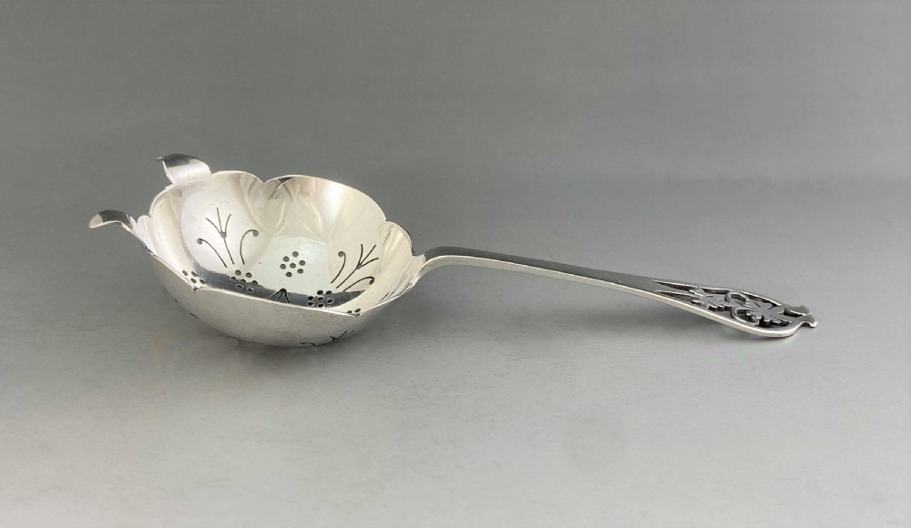 A silver tea strainer featuring a pierced bowl with geometric drainage holes and a decoratively cut-out flat handle. View III