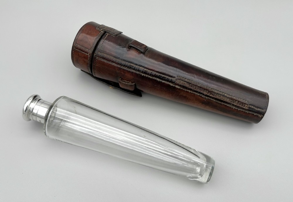 A conical clear glass hunting flask with a silver cap positioned next to its fitted dark brown leather saddle holster. View II