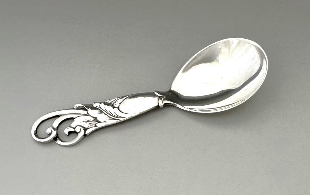 A Danish silver caddy spoon featuring a broad oval bowl and a handle decorated with carved foliage motifs and a scrolled terminal. View VI