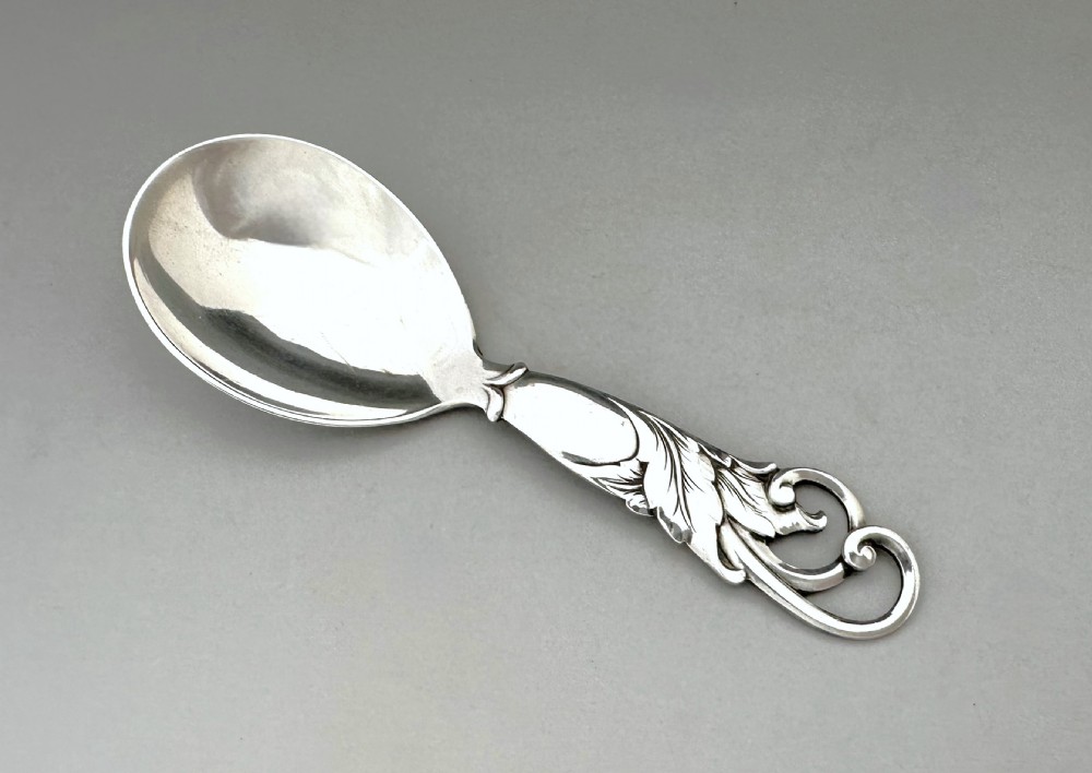 A Danish silver caddy spoon featuring a broad oval bowl and a handle decorated with carved foliage motifs and a scrolled terminal.