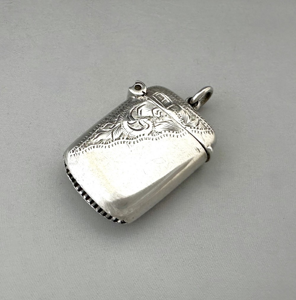 A small rectangular sterling silver vesta case featuring hand-engraved foliate decoration, a hinged lid, a suspension loop, and a serrated striker base.