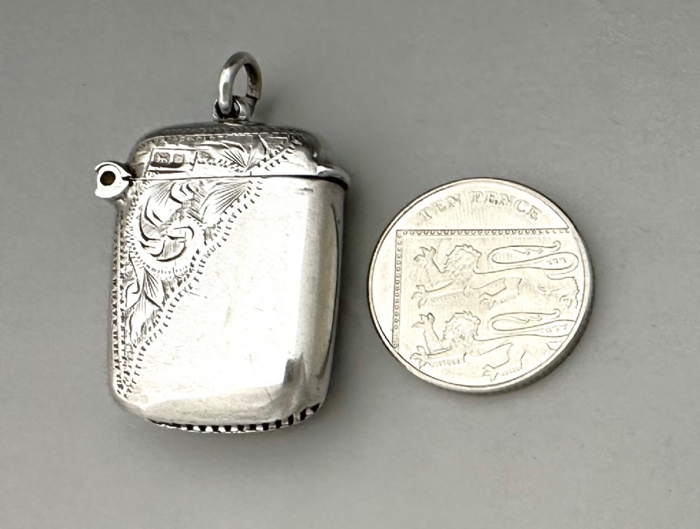 A small rectangular sterling silver vesta case featuring hand-engraved foliate decoration, a hinged lid, a suspension loop, and a serrated striker base. View VII