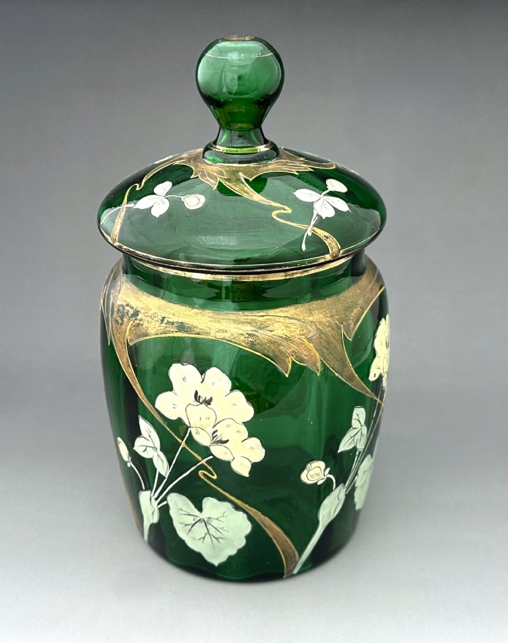 A green glass biscuit barrel featuring hand-painted white floral enamel and gold gilded leaf motifs with a matching glass lid.