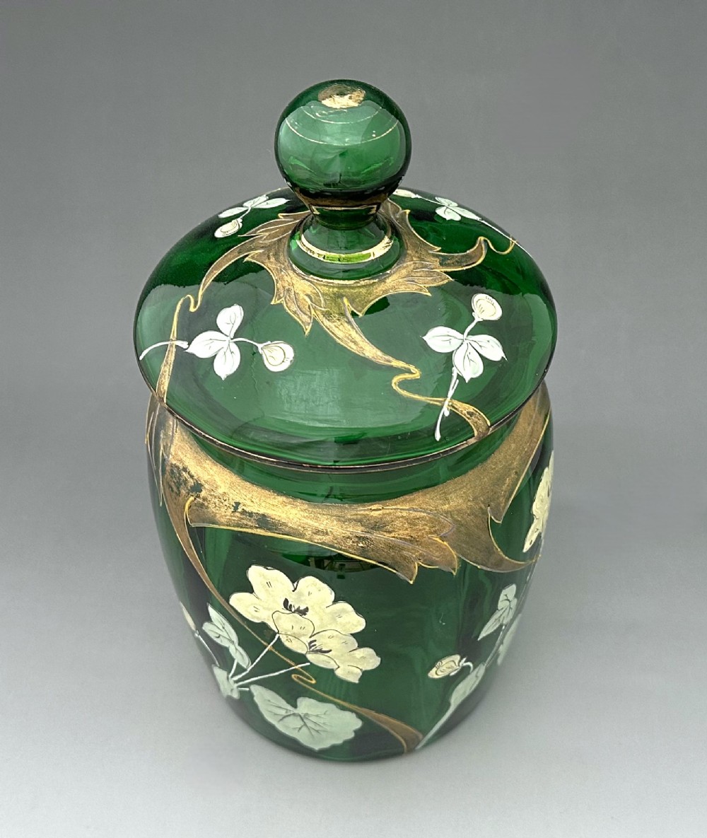 A green glass biscuit barrel featuring hand-painted white floral enamel and gold gilded leaf motifs with a matching glass lid. View II
