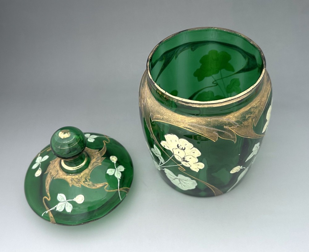A green glass biscuit barrel featuring hand-painted white floral enamel and gold gilded leaf motifs with a matching glass lid. View V