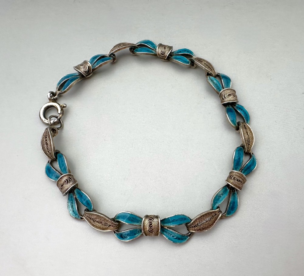 A silver filigree link bracelet featuring bow-shaped segments filled with turquoise blue enamel and secured with a bolt ring clasp.
