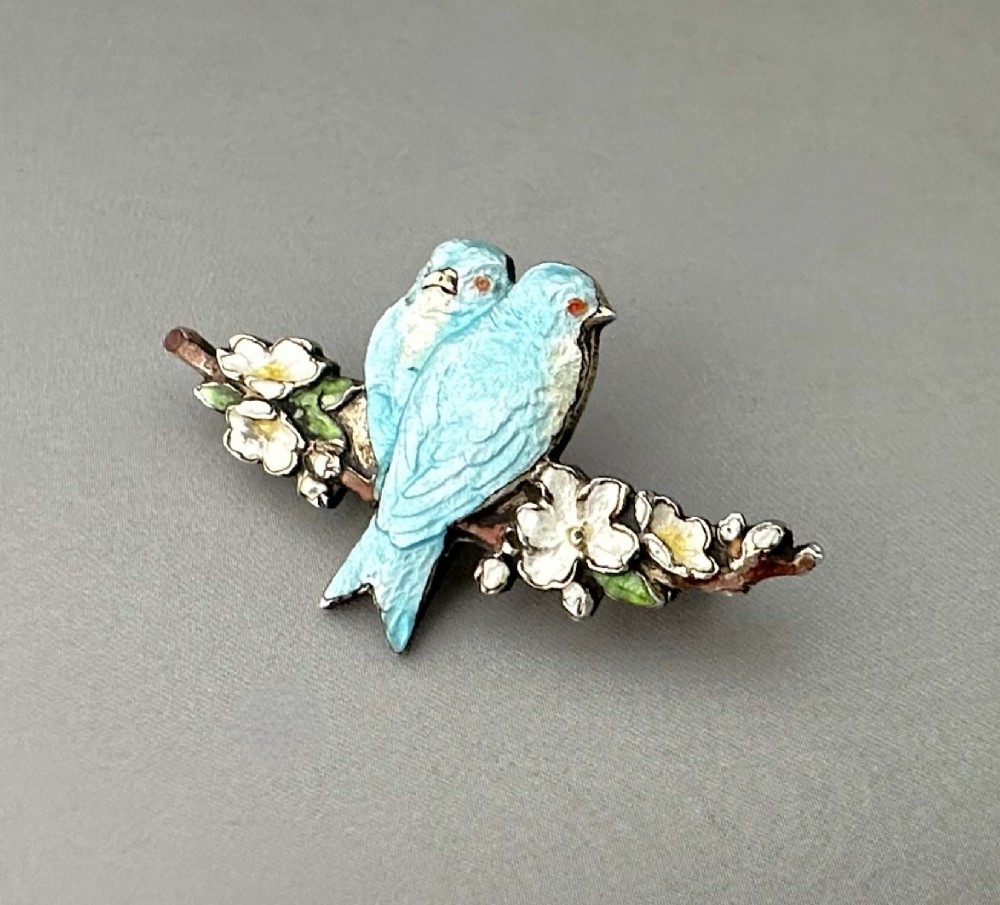 A silver-gilt brooch depicting two light blue enamel birds perched on a flowering branch with white enamel blossoms.