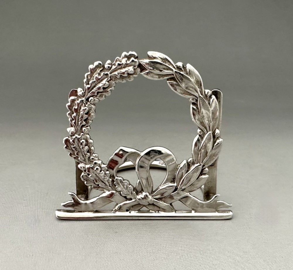 A pair of sterling silver menu card holders shaped as laurel and oak leaf wreaths tied with a ribbon bow on a flat base with rear support struts. View V