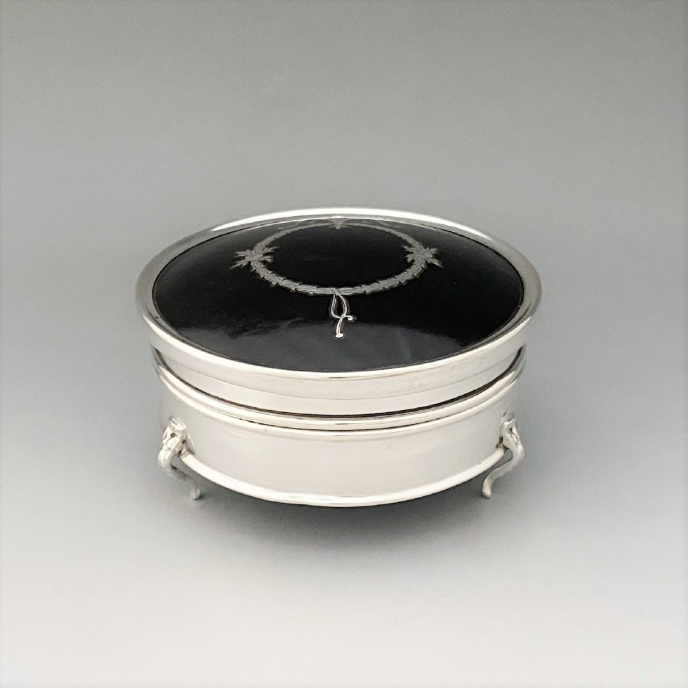 A circular sterling silver trinket box resting on four cabriole legs featuring a dark tortoiseshell lid inlaid with a silver floral garland motif.