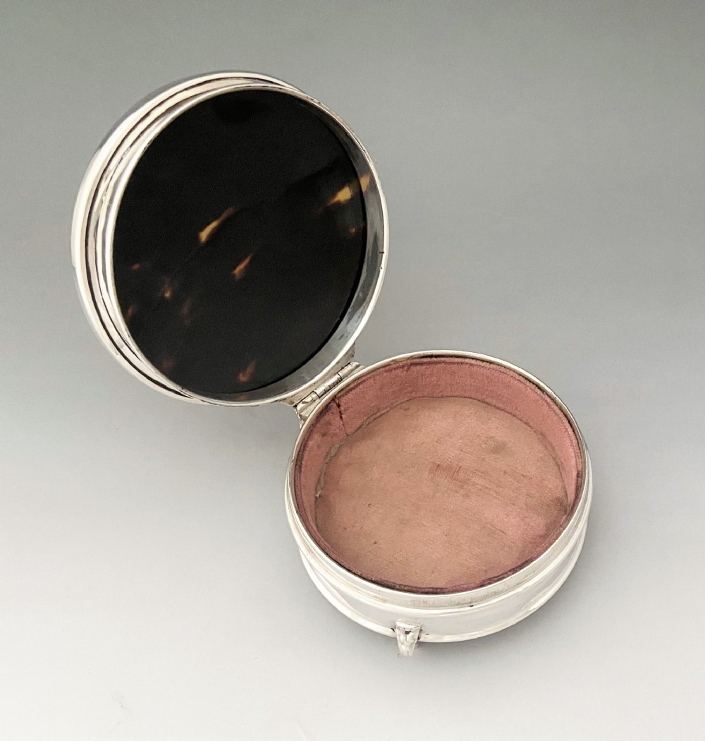 A circular sterling silver trinket box resting on four cabriole legs featuring a dark tortoiseshell lid inlaid with a silver floral garland motif. View V