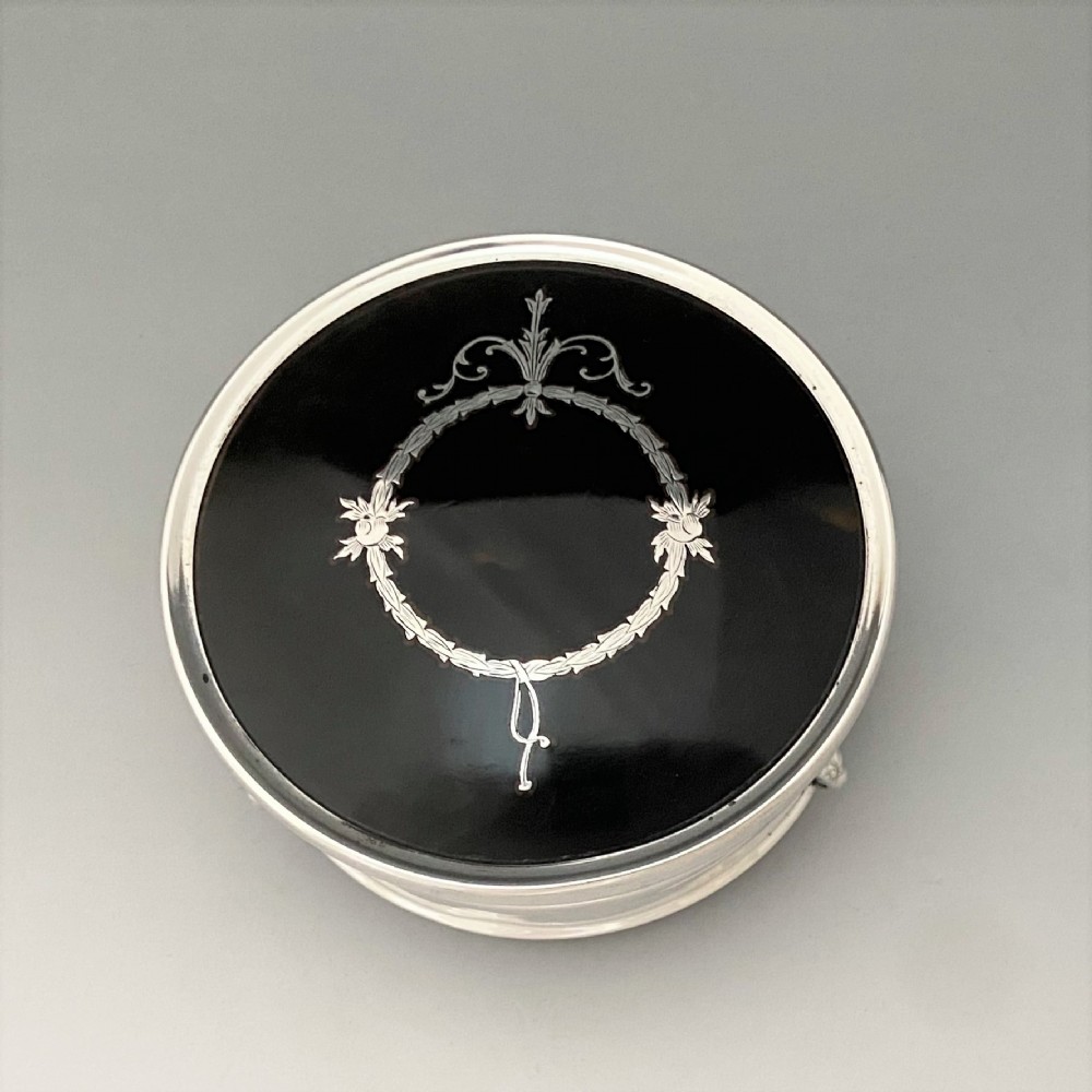 A circular sterling silver trinket box resting on four cabriole legs featuring a dark tortoiseshell lid inlaid with a silver floral garland motif. View VI