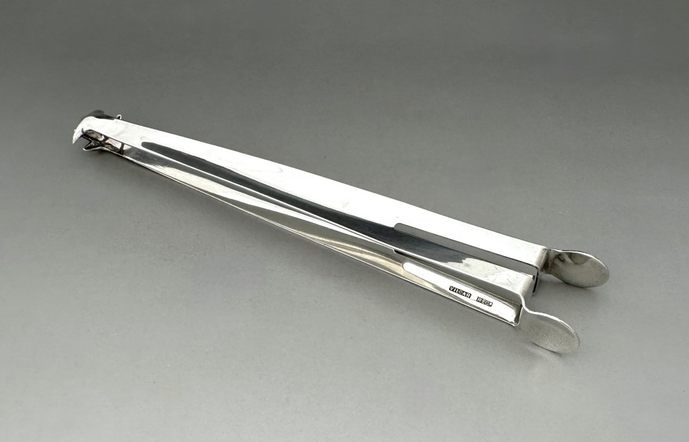 A pair of polished silver Edwardian bar tongs featuring slender arms with mechanical claw gripping ends and flat finger pads.