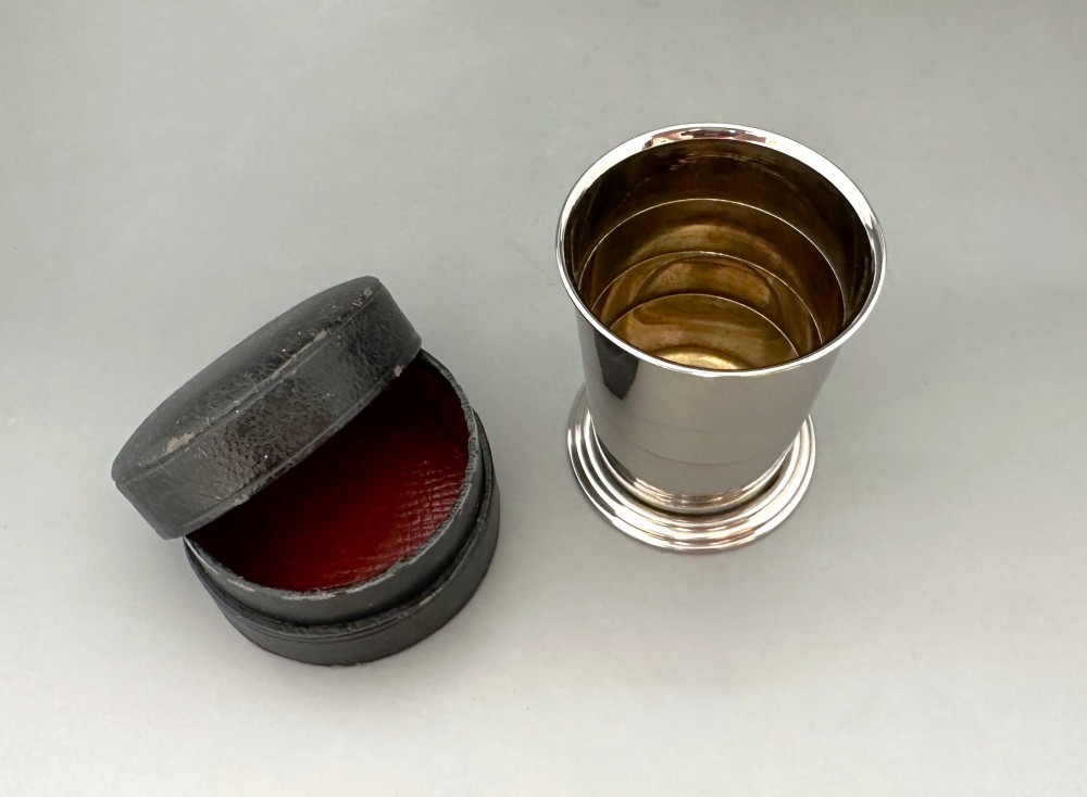 A silver-plated collapsible cup composed of concentric metal rings standing fully extended next to its open dark leather storage case. View II