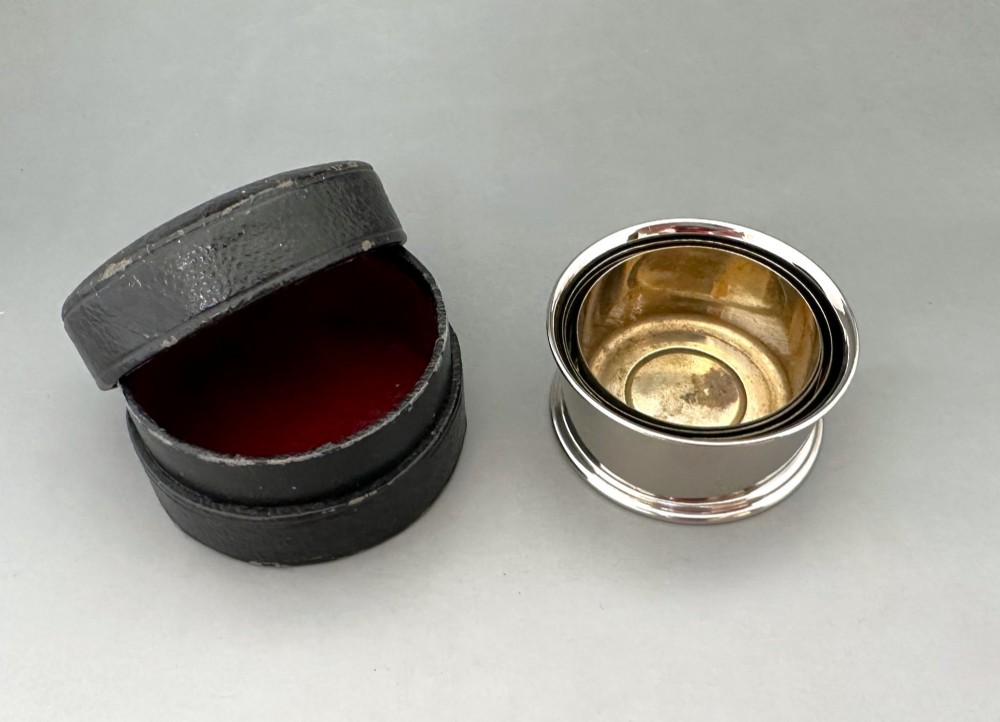 A silver-plated collapsible cup composed of concentric metal rings standing fully extended next to its open dark leather storage case. View III
