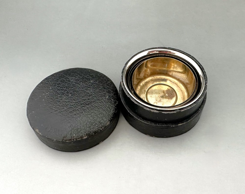 A silver-plated collapsible cup composed of concentric metal rings standing fully extended next to its open dark leather storage case. View IV