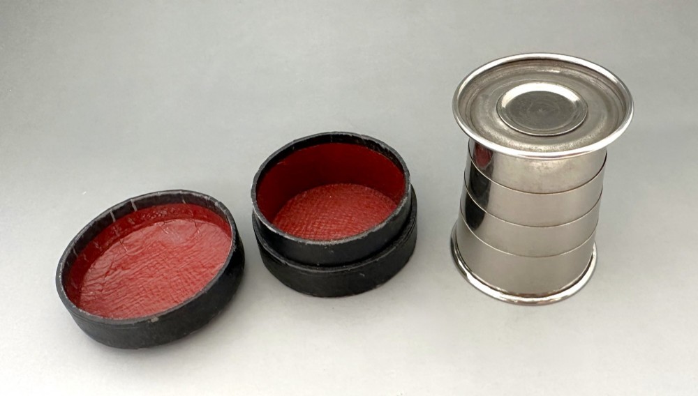 A silver-plated collapsible cup composed of concentric metal rings standing fully extended next to its open dark leather storage case. View V