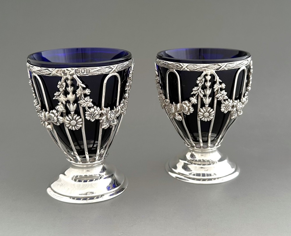 A pair of silver wirework condiment tubs featuring cast floral garlands and cobalt blue glass liners on round pedestal bases.