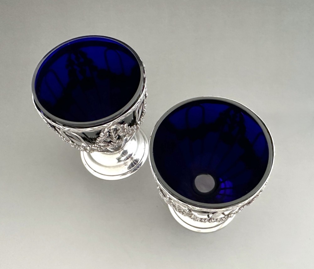 A pair of silver wirework condiment tubs featuring cast floral garlands and cobalt blue glass liners on round pedestal bases. View IV