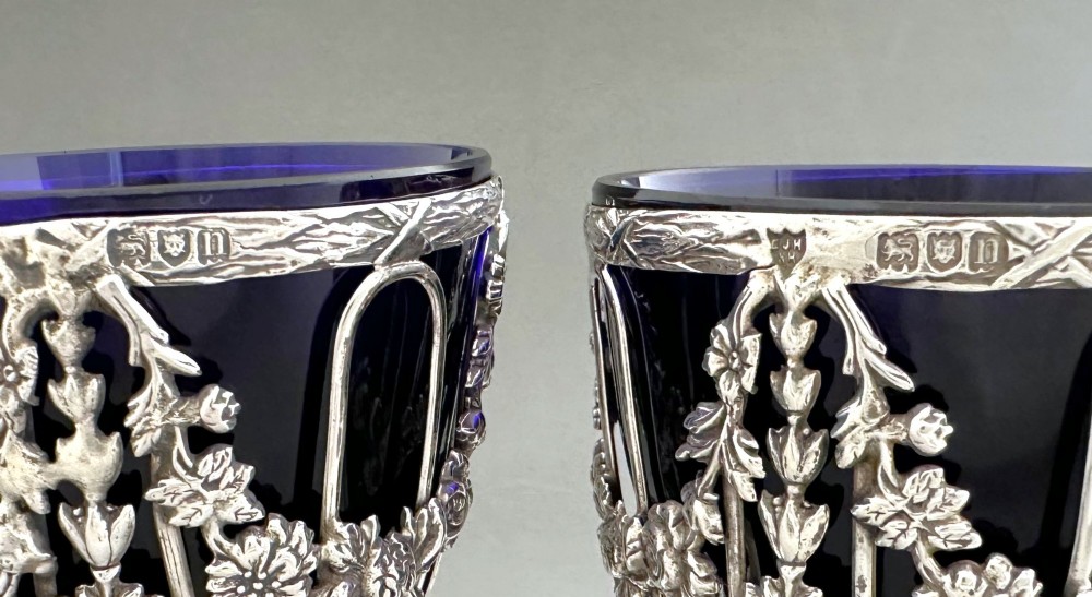 A pair of silver wirework condiment tubs featuring cast floral garlands and cobalt blue glass liners on round pedestal bases. View VIII