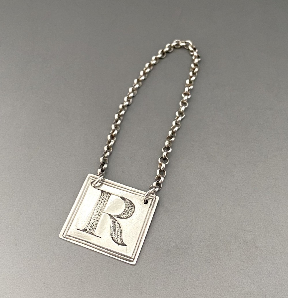 A square silver decanter label with a moulded border, engraved with the letter R and attached to a delicate silver chain.
