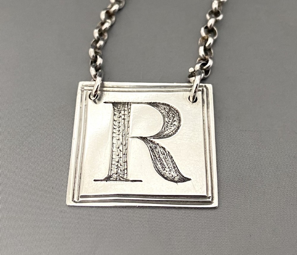 A square silver decanter label with a moulded border, engraved with the letter R and attached to a delicate silver chain. View II