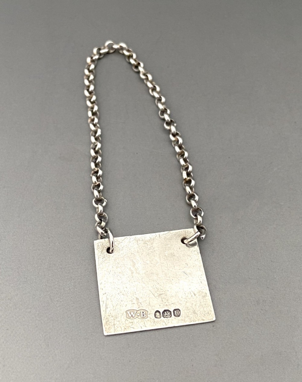 A square silver decanter label with a moulded border, engraved with the letter R and attached to a delicate silver chain. View III