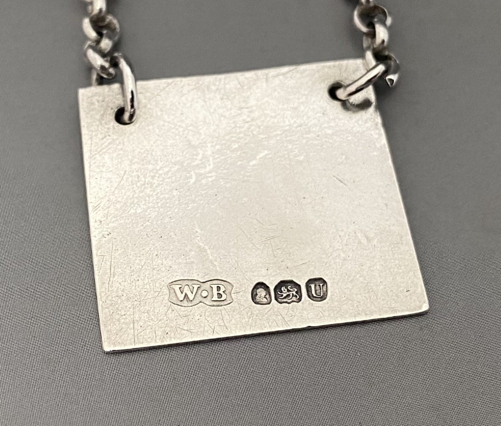 A square silver decanter label with a moulded border, engraved with the letter R and attached to a delicate silver chain. View IV