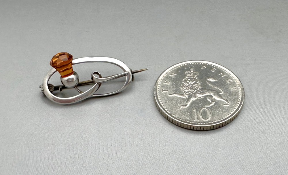 An Art Nouveau silver brooch featuring a stylised oval frame with a central thistle motif topped with a faceted amber-coloured glass stone. View VI