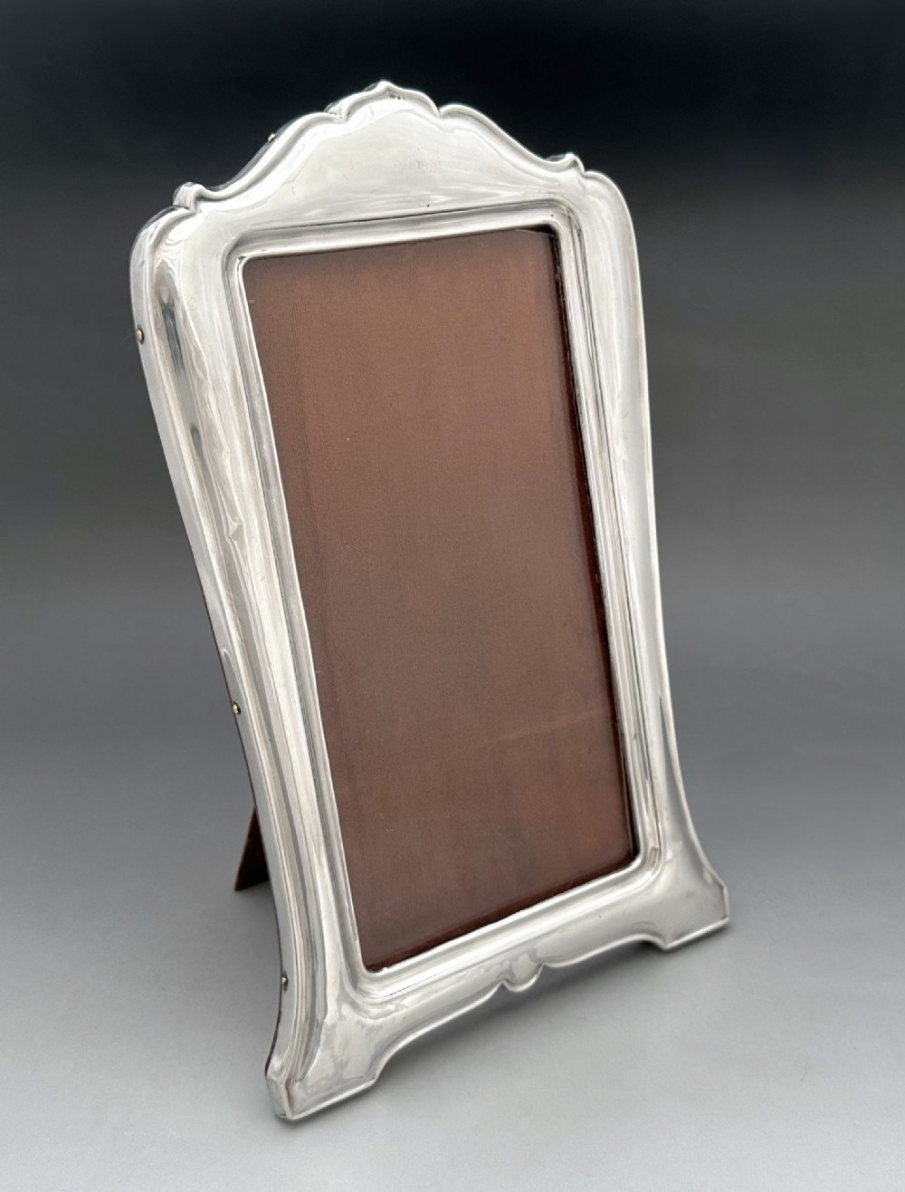 A George V polished silver picture frame with a shaped arched top and a dark wooden easel backing.