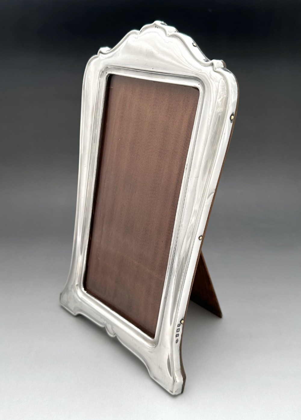 A George V polished silver picture frame with a shaped arched top and a dark wooden easel backing. View II