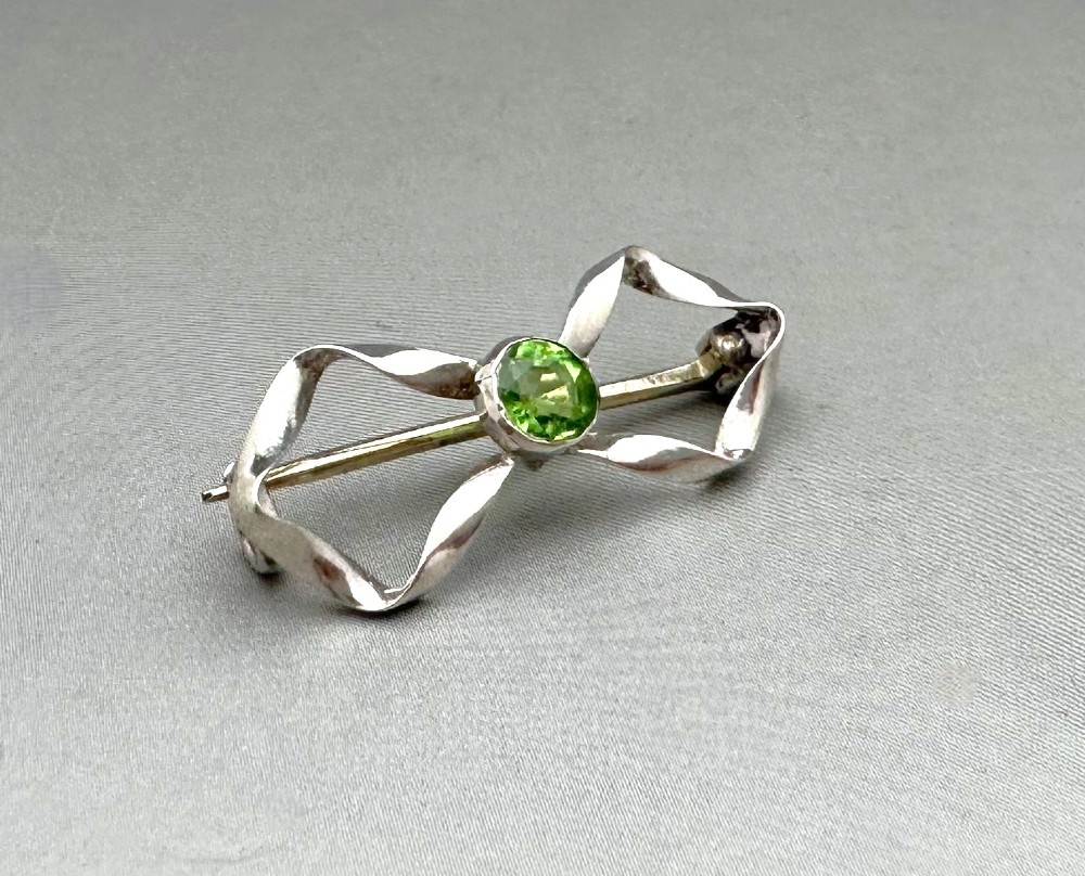 An Edwardian sterling silver brooch styled as a ribbon bow with a faceted green peridot stone set in the centre.