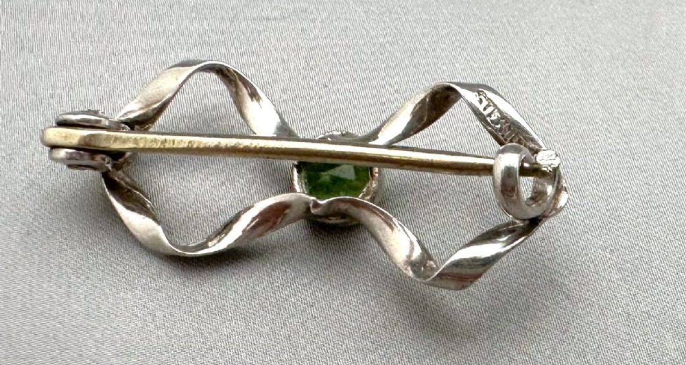An Edwardian sterling silver brooch styled as a ribbon bow with a faceted green peridot stone set in the centre. View III