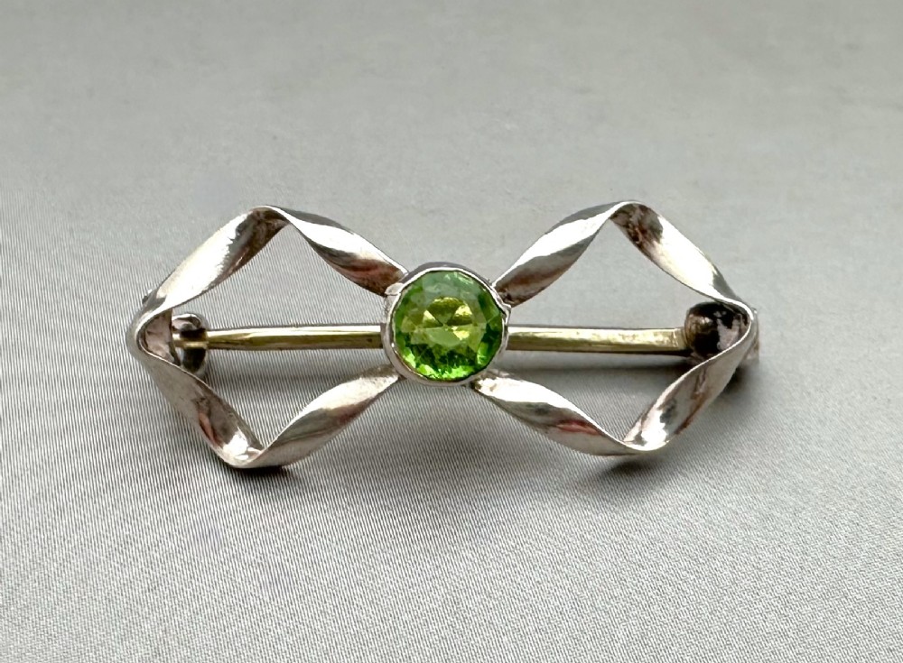 An Edwardian sterling silver brooch styled as a ribbon bow with a faceted green peridot stone set in the centre. View IV