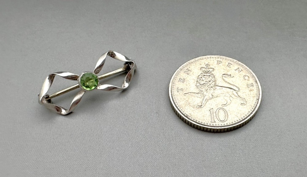 An Edwardian sterling silver brooch styled as a ribbon bow with a faceted green peridot stone set in the centre. View V
