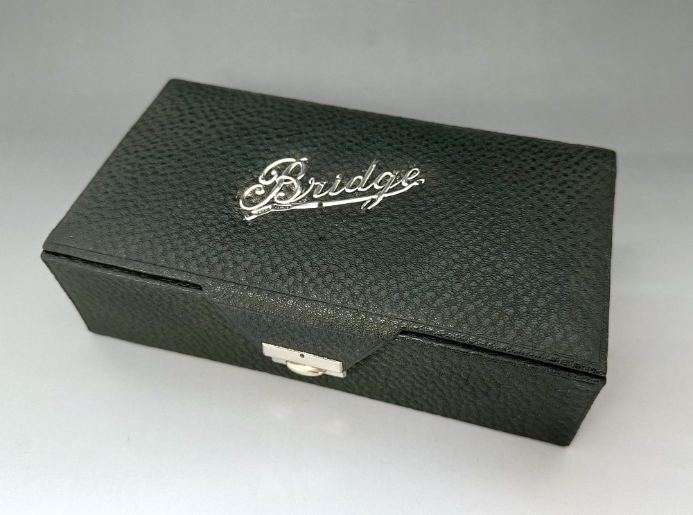 A dark green pebbled leather rectangular bridge box featuring applied silver script spelling Bridge on the top cover and a silver clasp closure.