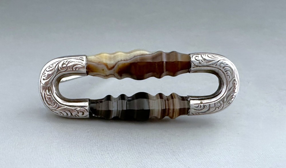 An elongated Victorian silver brooch featuring hand-engraved end mounts and faceted banded brown agate stones. View V